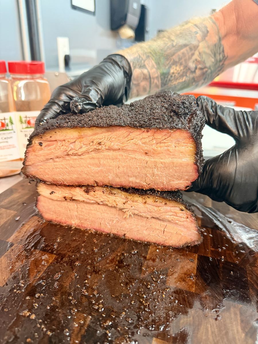 Pitmaster's gloved hands holding a thick cut of smoked brisket with a dark peppery bark