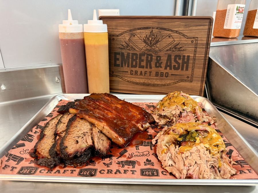 Tray of spare ribs, sliced brisket and pulled pork in front of an Ember & Ash Craft BBQ wooden sign